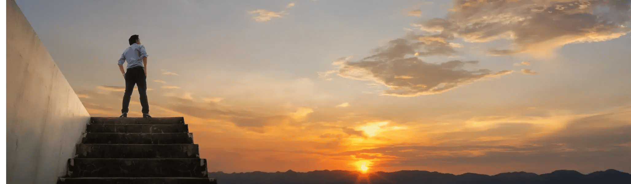 階段を上りきった先で、夕日に染まる空と山並みを静かに見つめるビジネスパーソンの後ろ姿_メインビジュアル_pc